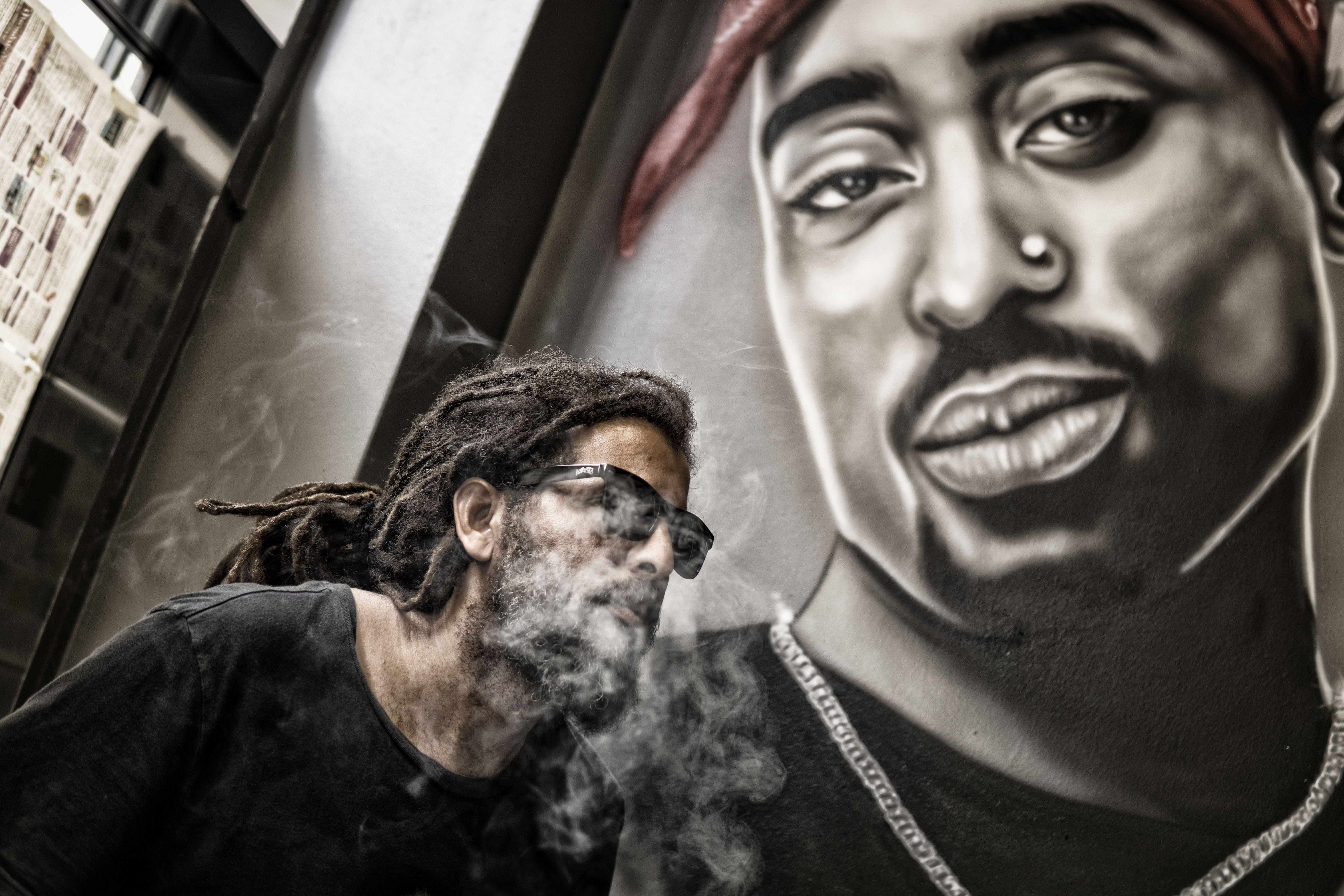 A man smoking in front of a mural featuring a hip hop artist, blending urban style and art.
