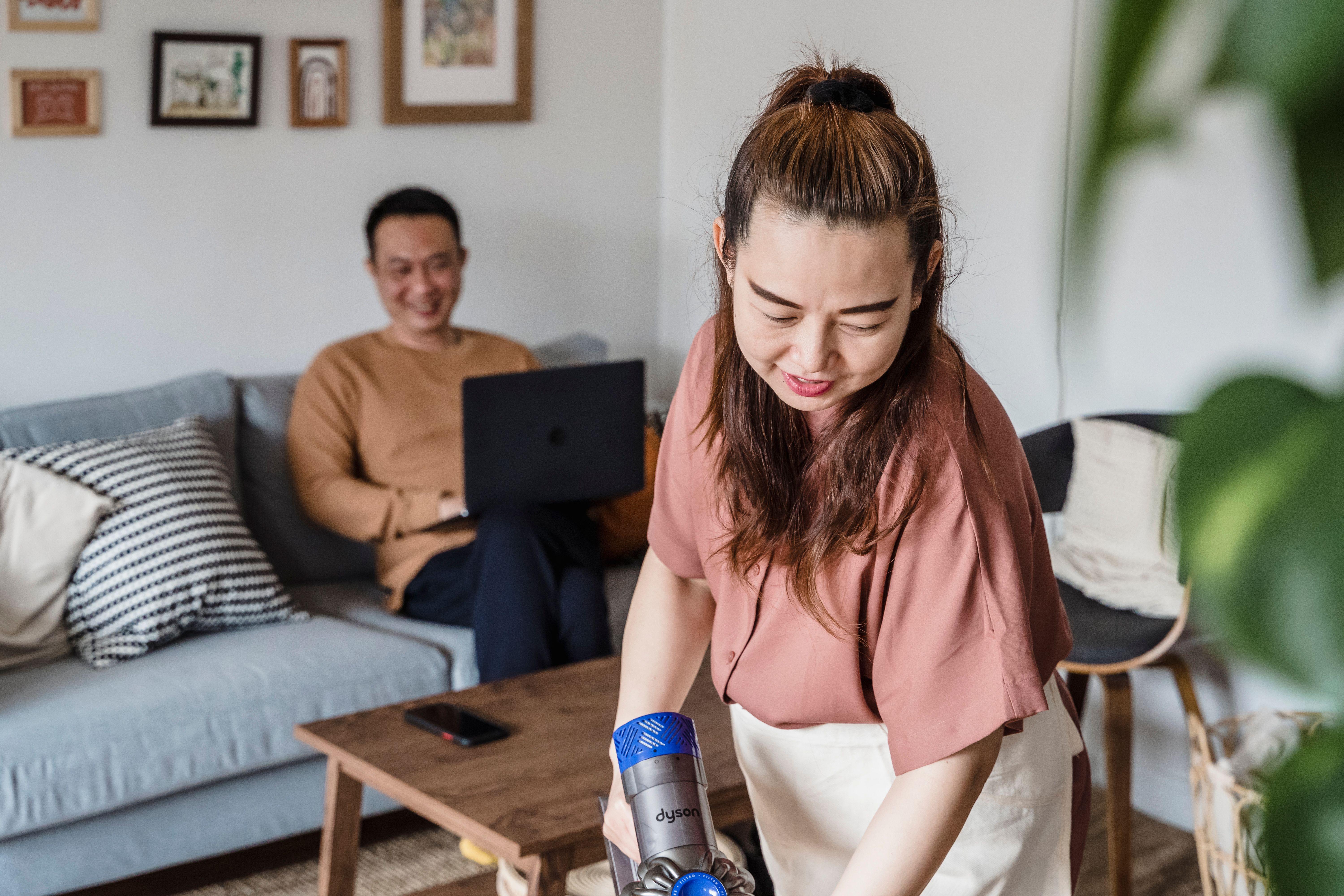 A woman smiling while vacuuming a living room, with a man working on a laptop in the background.