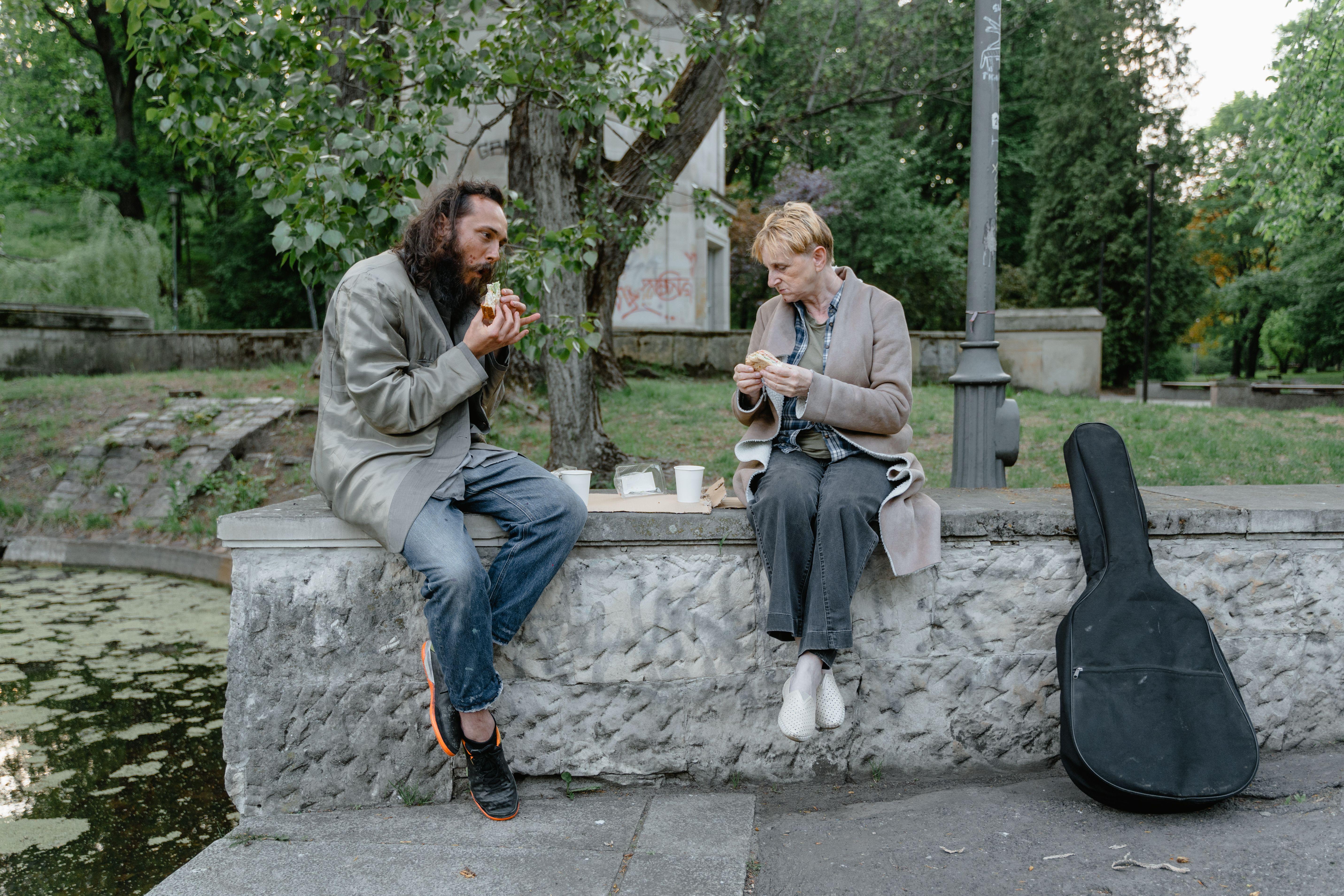 Two individuals sharing a meal outdoors on a stone bench, highlighting companionship and resilience.