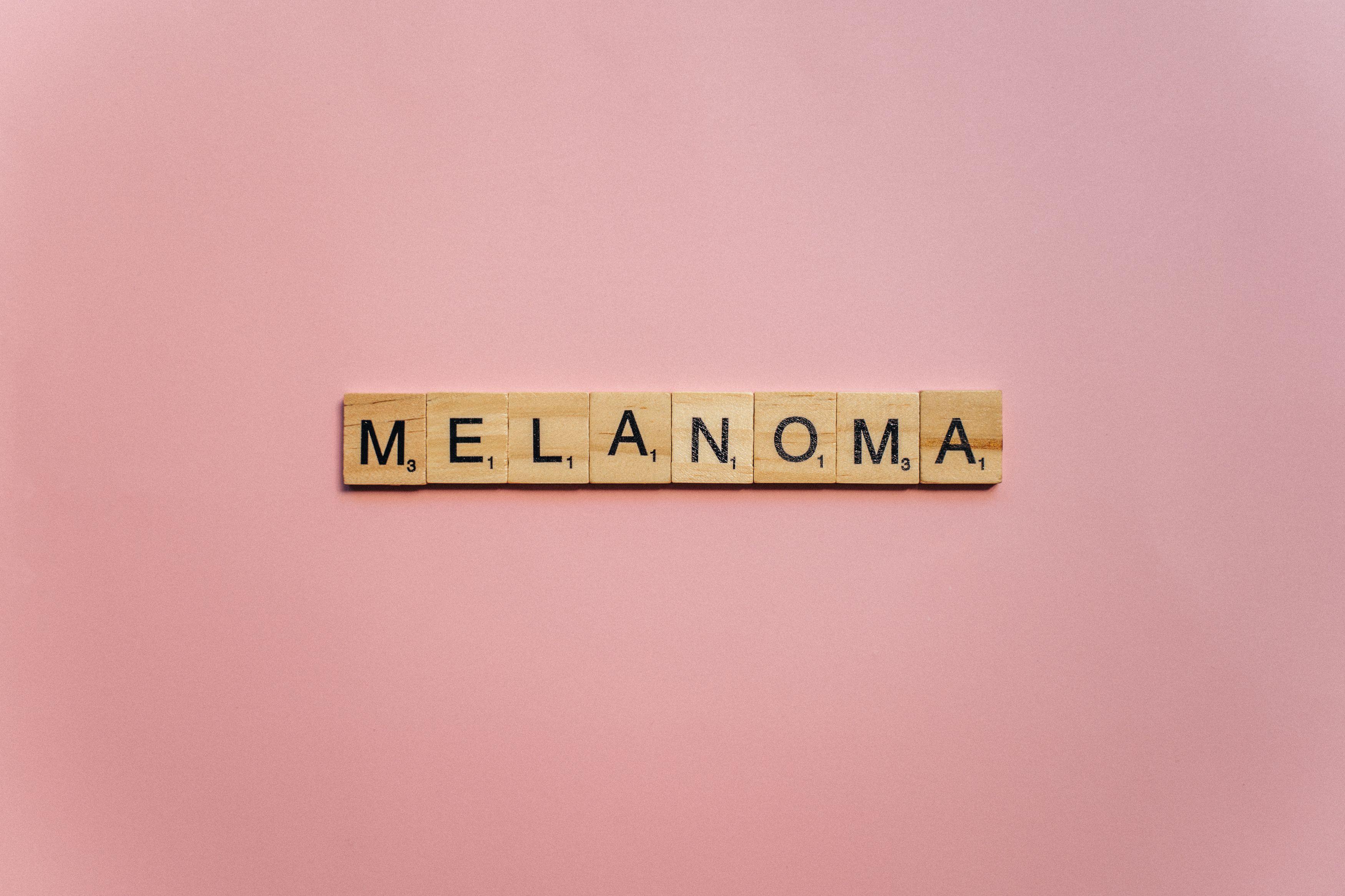 Wooden Scrabble tiles spelling 'Melanoma' on a soft pink background, symbolizing awareness.