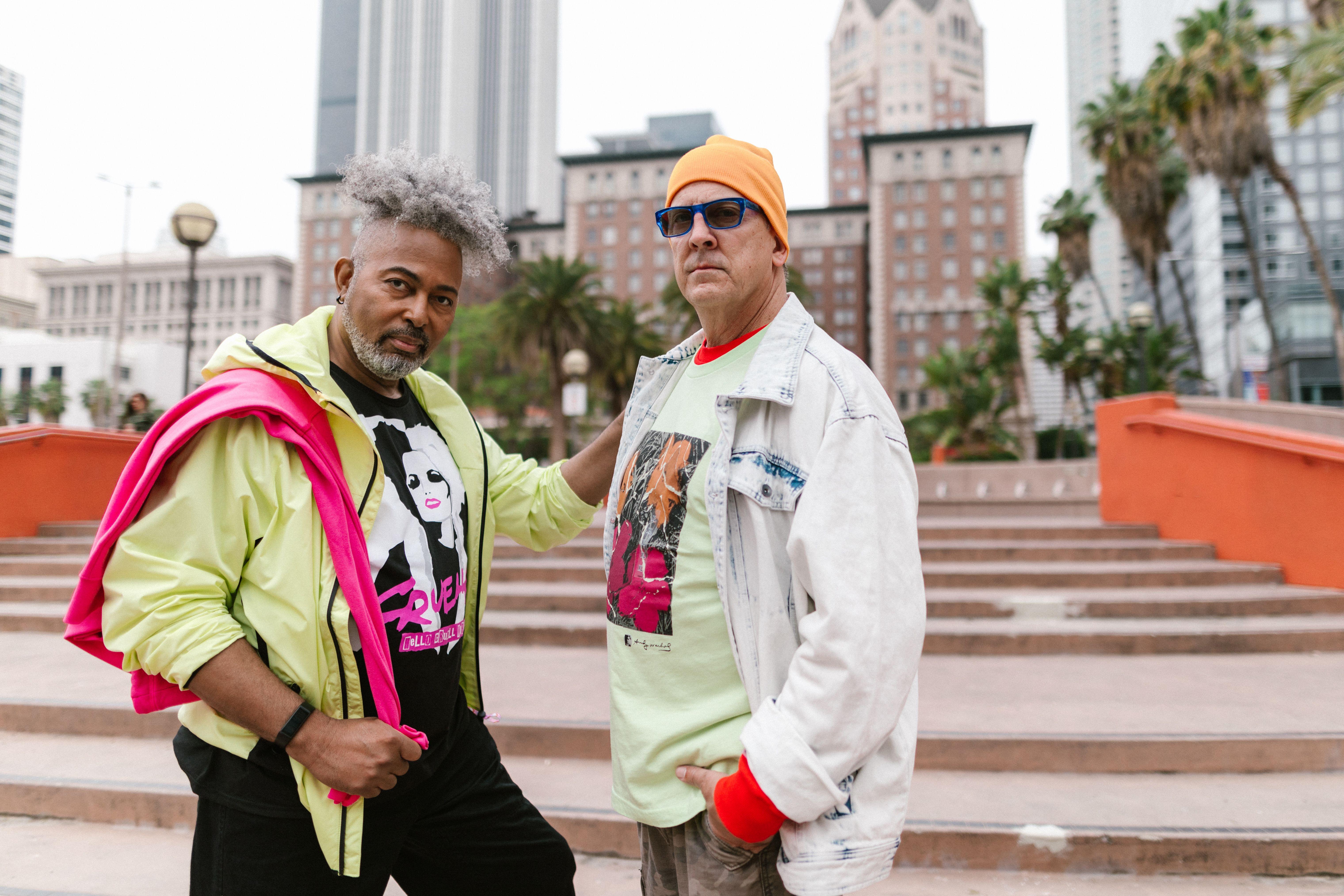 Two fashionable men pose confidently in a vibrant urban setting with city skyscrapers as backdrop.