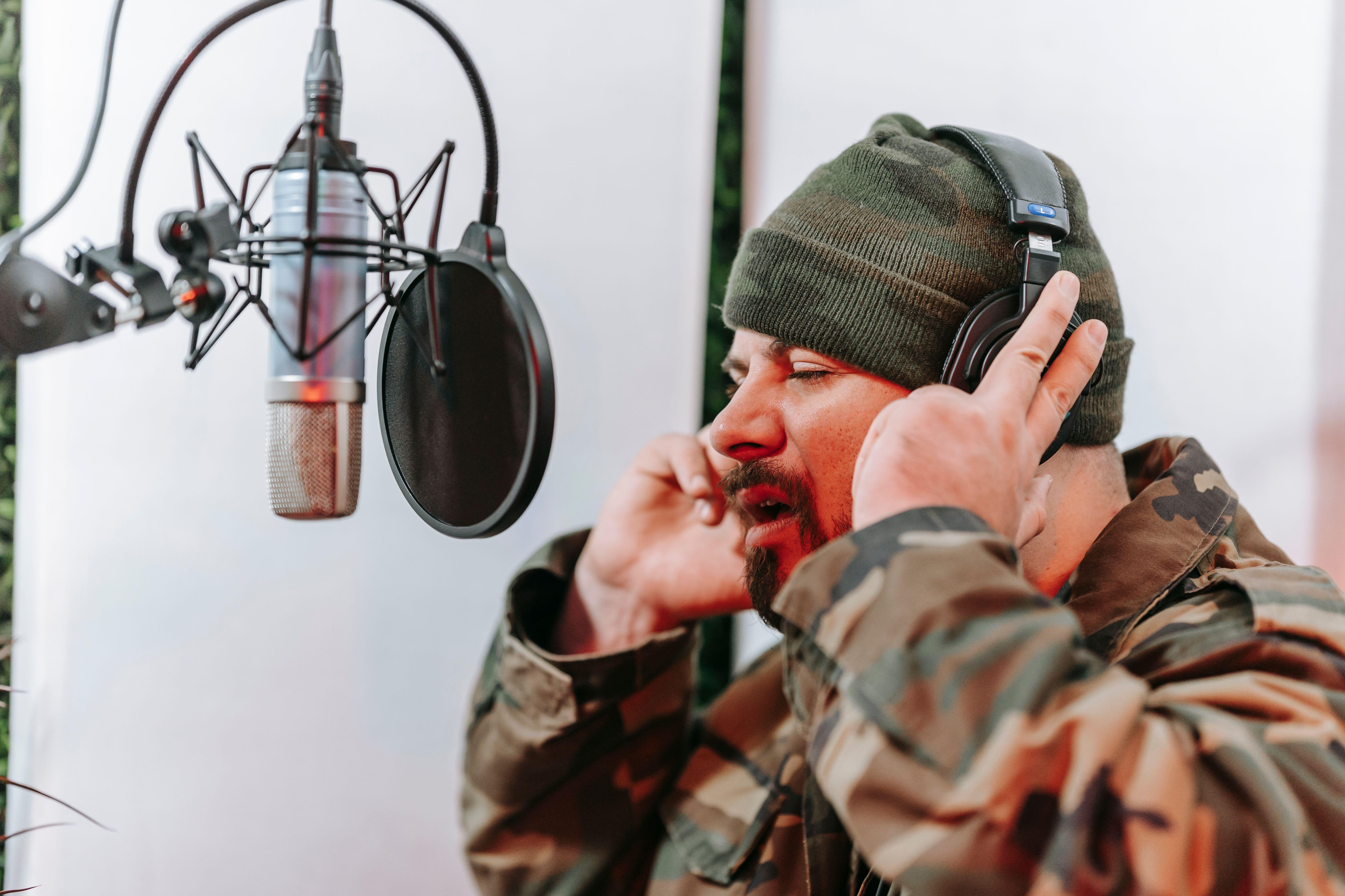Man in a camouflage jacket singing into a microphone in a studio setting with headphones on, focused and expressive.