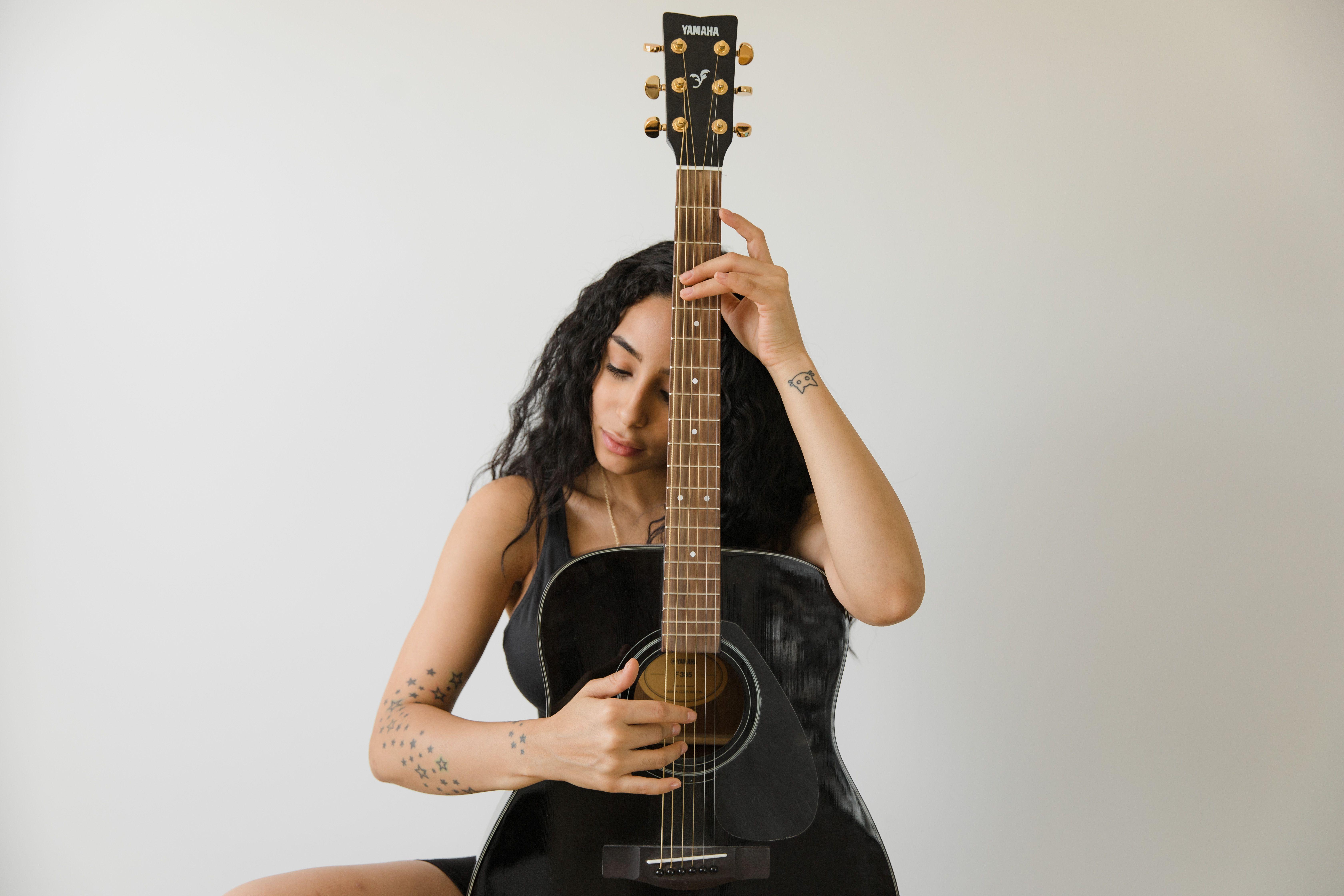 Woman with curly hair plays a black acoustic guitar indoors, focusing on her music.