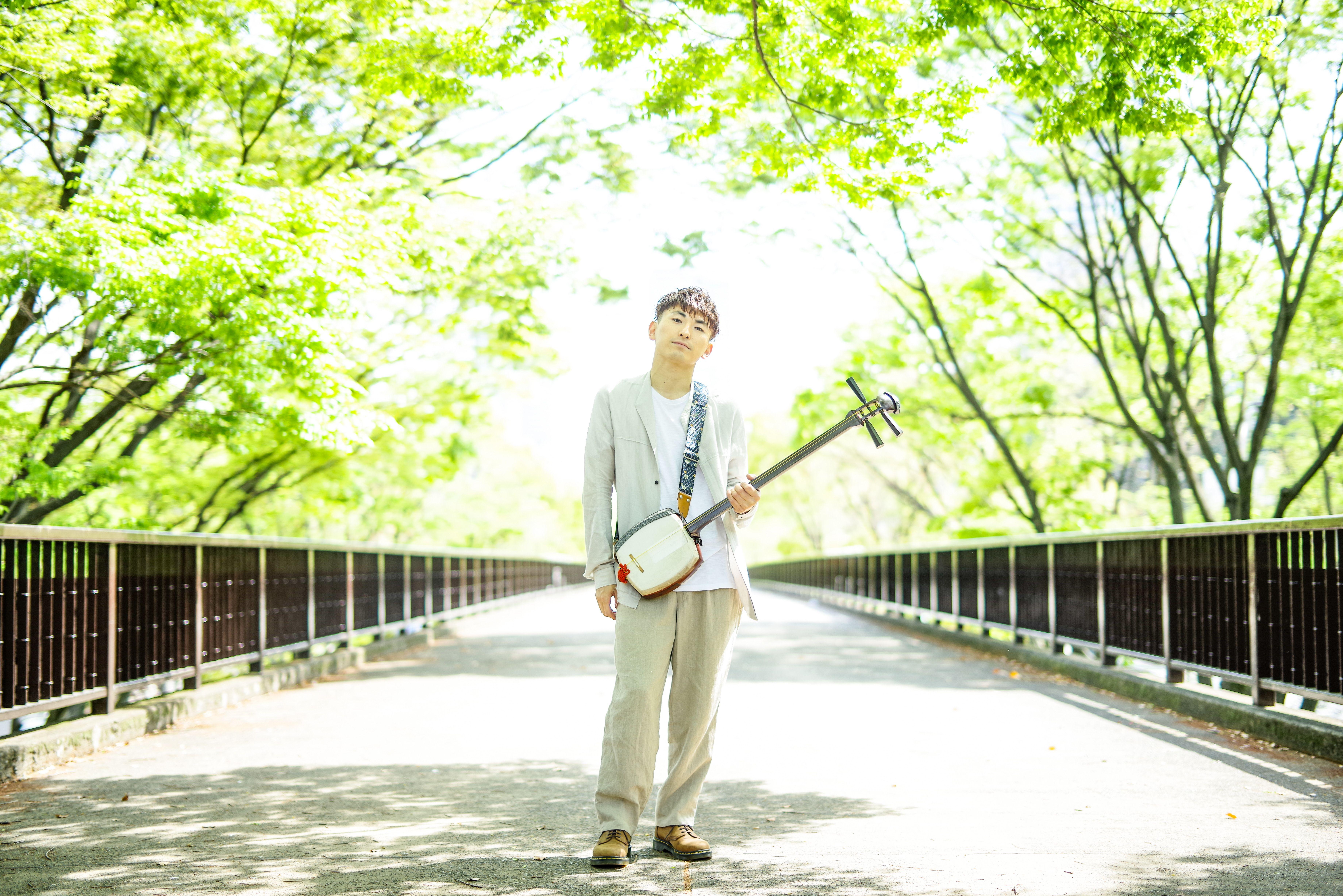 An Asian man standing on a sunlit bridge, holding a shamisen, surrounded by vibrant green trees.