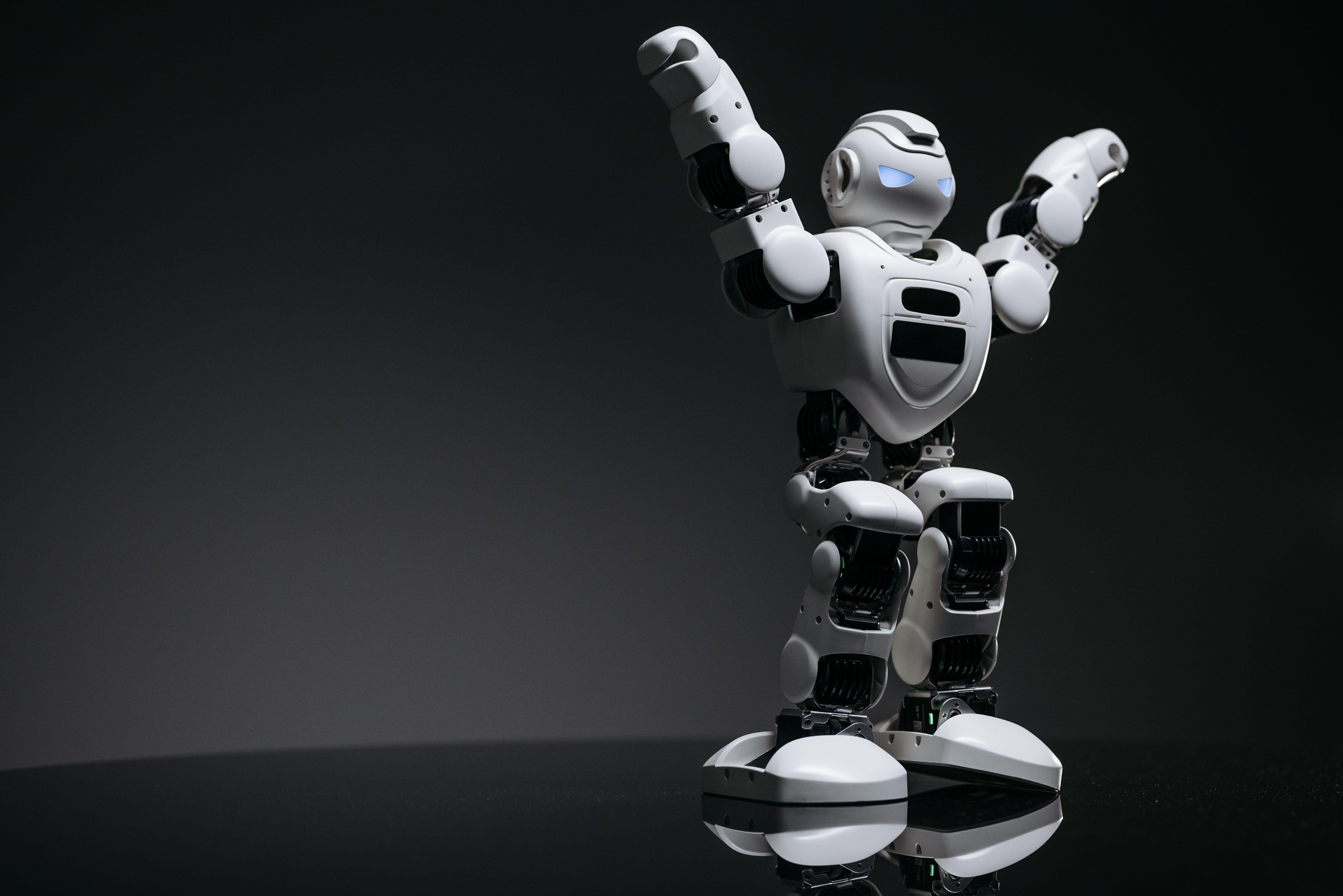 Close-up studio shot of a white robot toy with LED eyes raised in victory on a gray background.