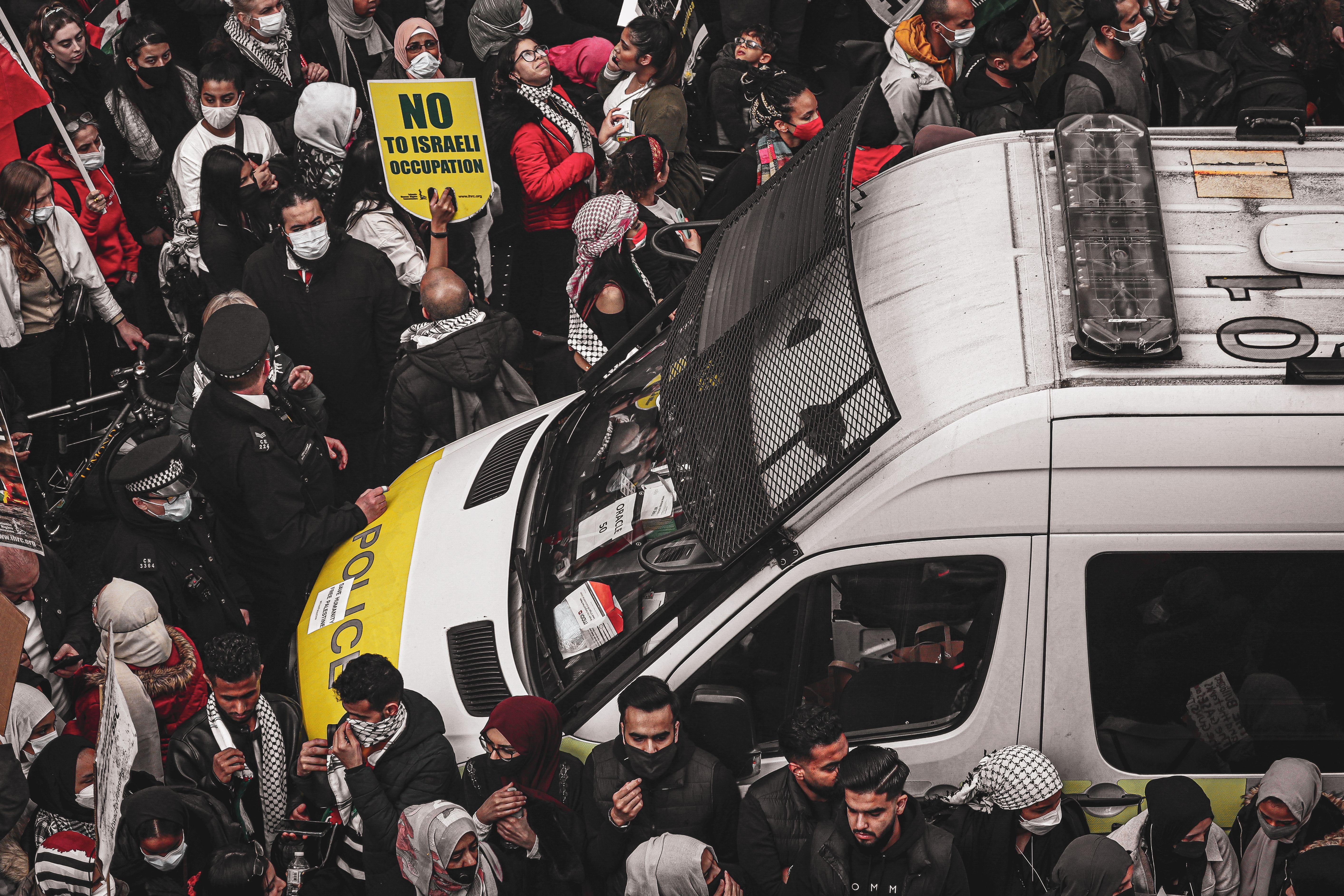 A crowd of protesters surrounding a police van holding signs during a demonstration against Israeli occupation.