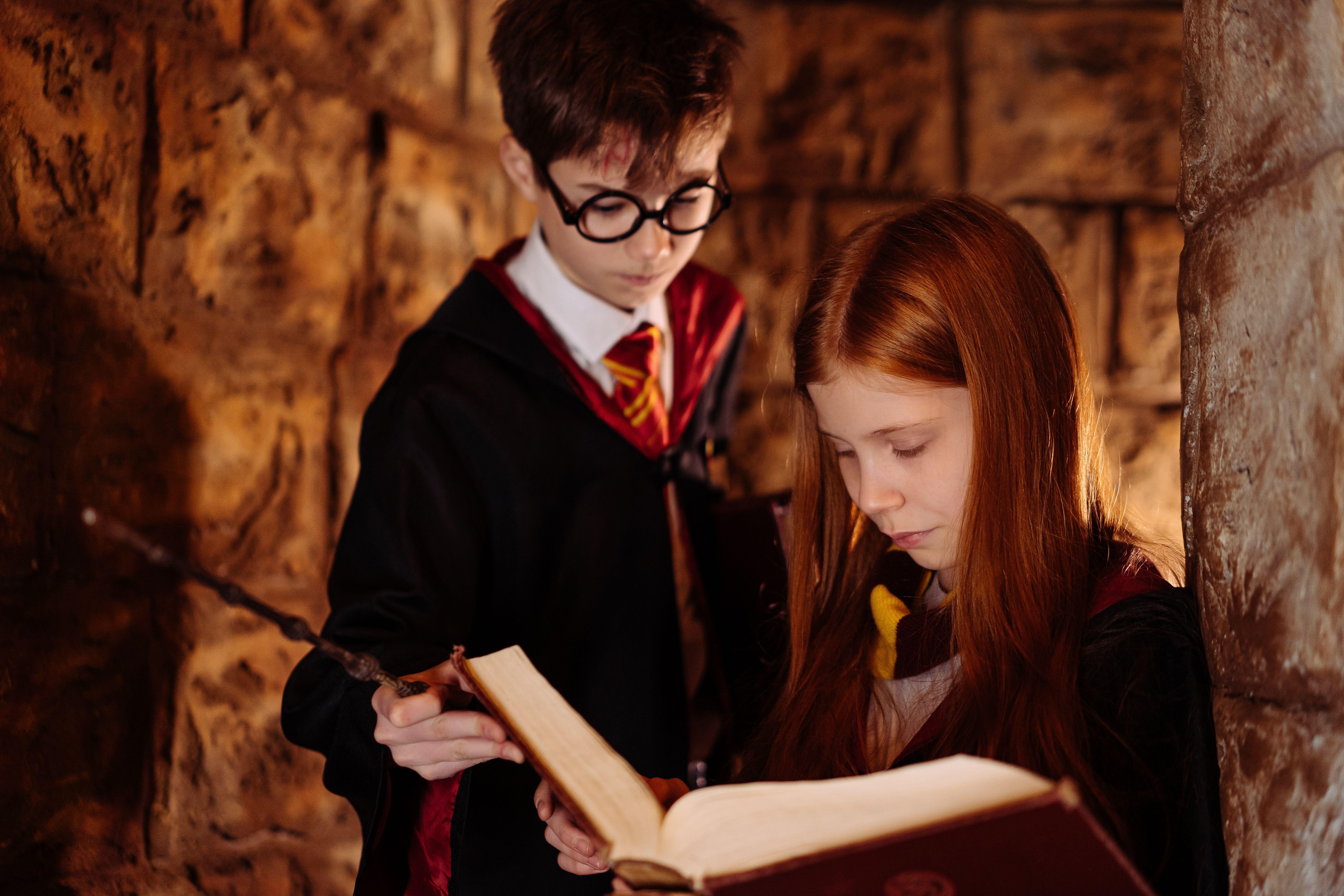 Two children in wizard costumes reading a spellbook in a dimly lit stone room.