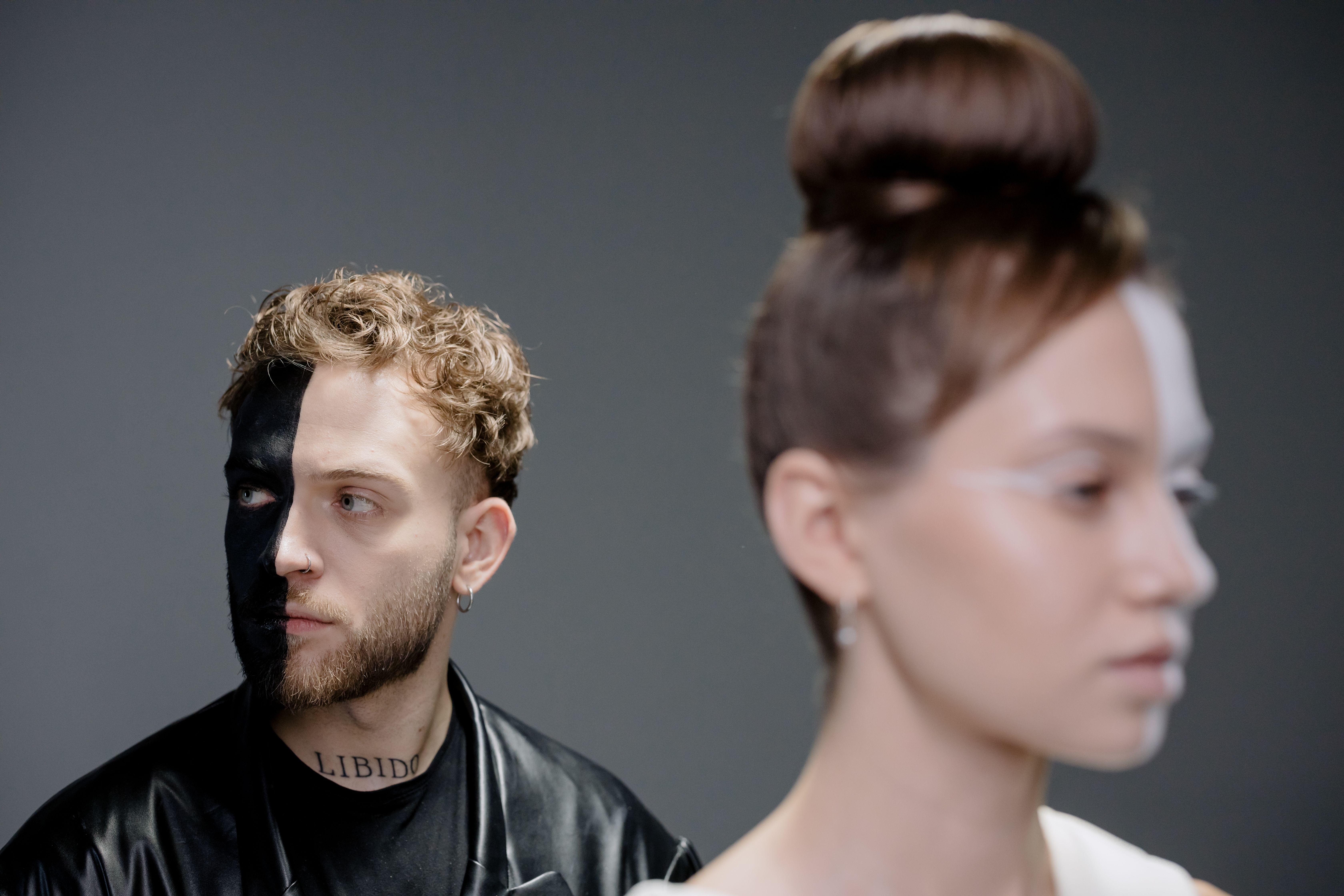 Artistic portrait featuring two models with creative half-face paint against a gray background.