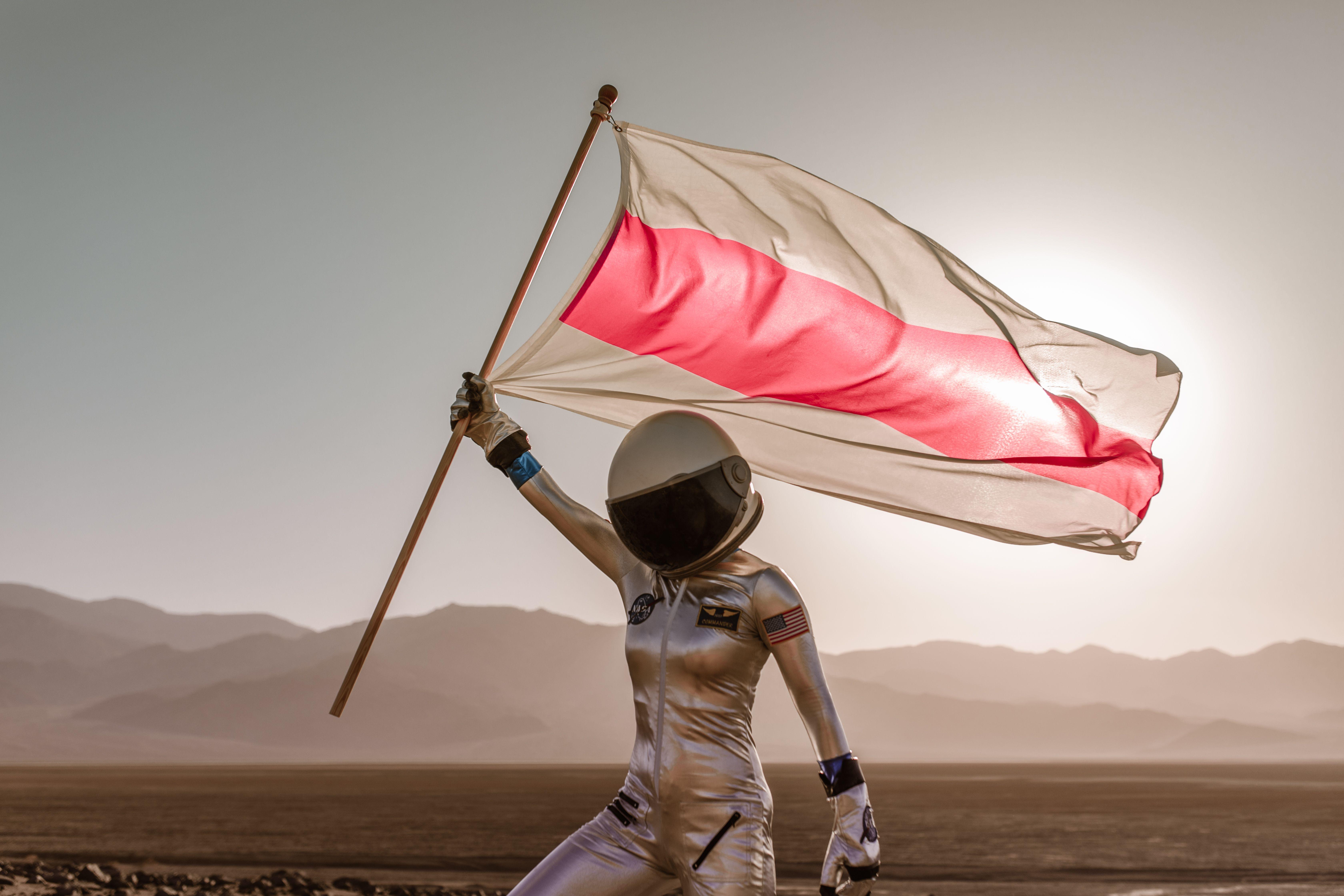 Astronaut in spacesuit holding a red and white flag in a desert setting with mountains in the background.