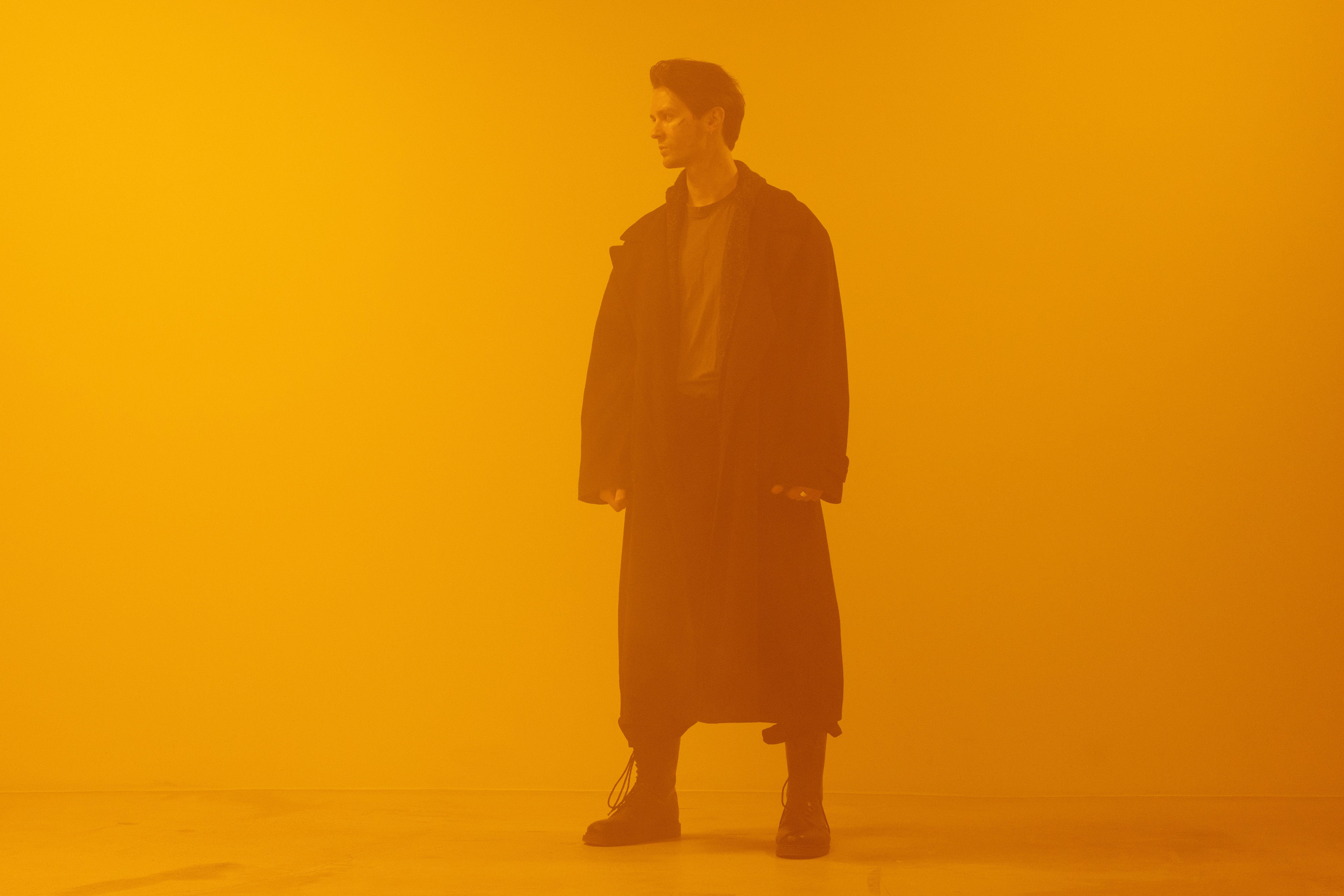Man in black coat standing against an orange background, embodying a cyberpunk retro-futuristic style.