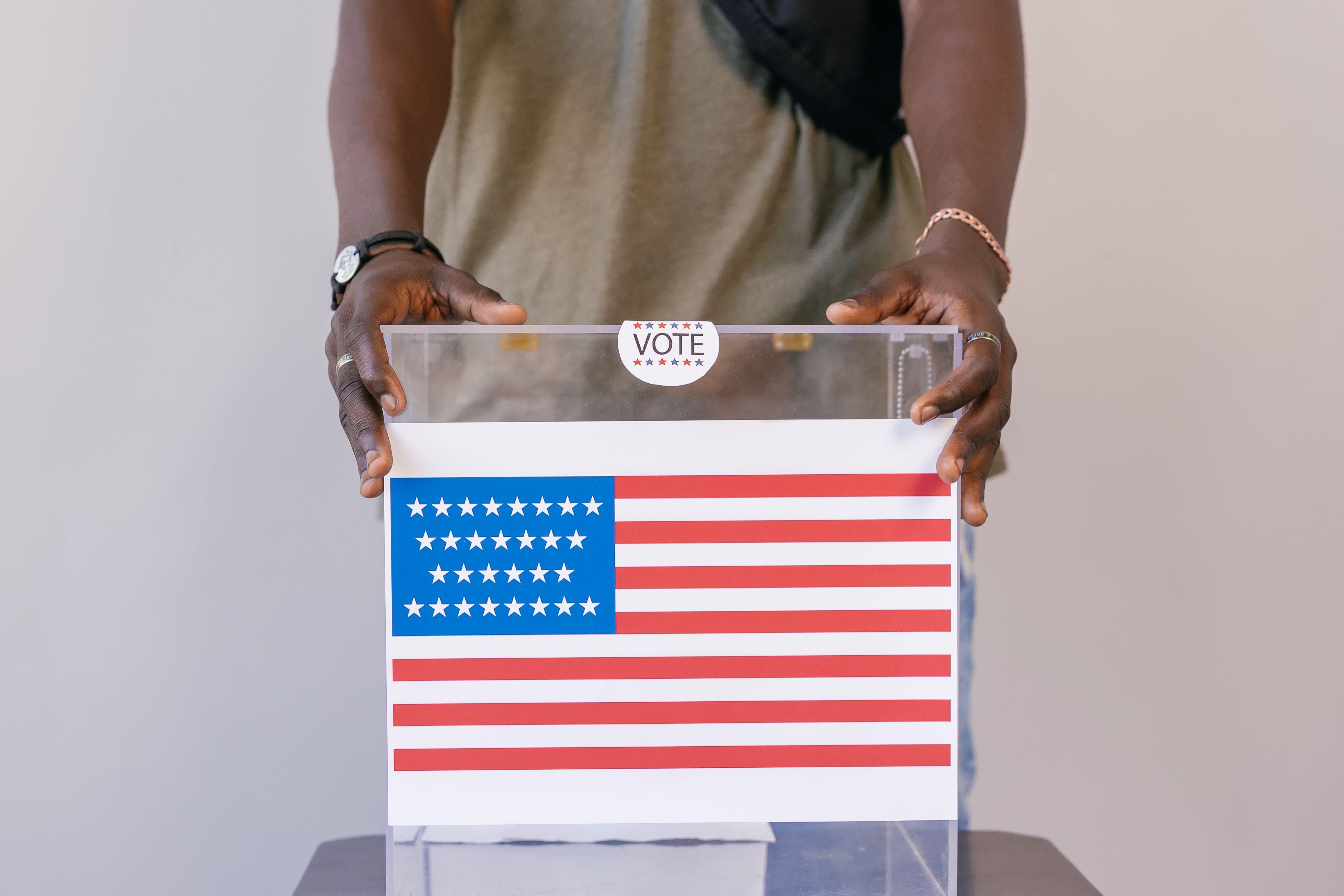 Close-up of a person placing a vote in a transparent ballot box with an American flag print.