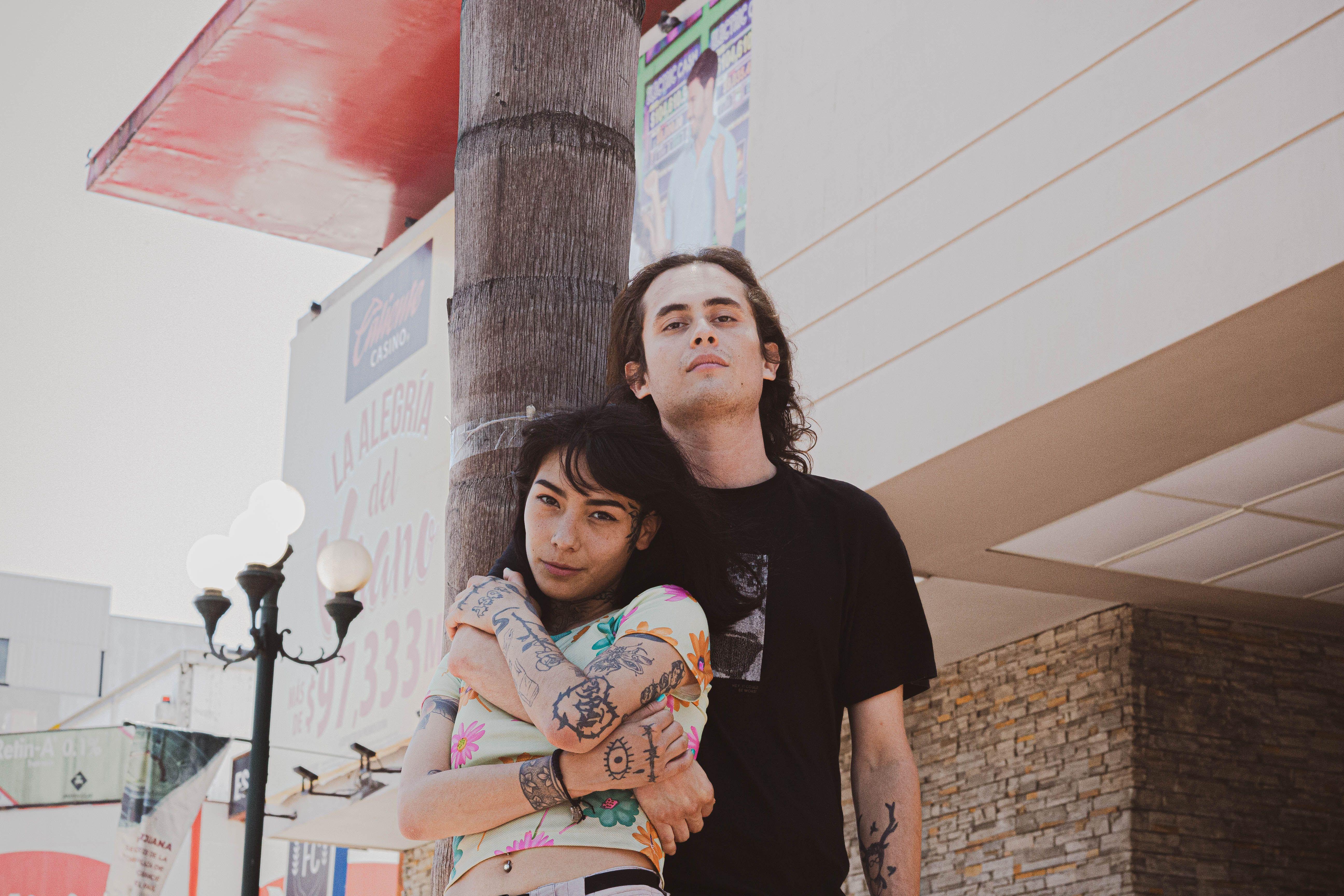 A stylish young couple embraces in an urban outdoor setting, showcasing tattoos and streetwear fashion.