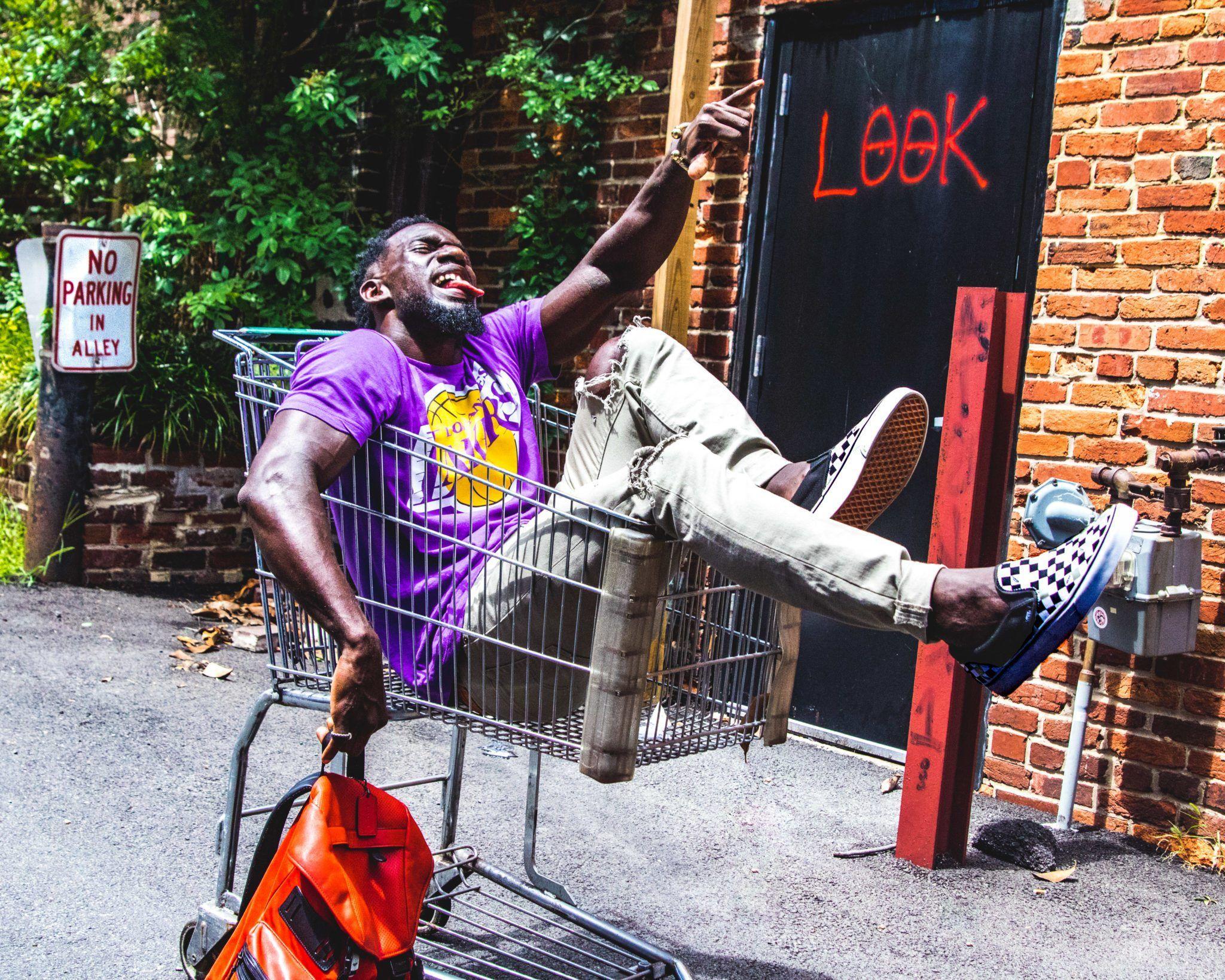 A joyful man posing in a shopping cart in an urban alley, embracing carefree fun.