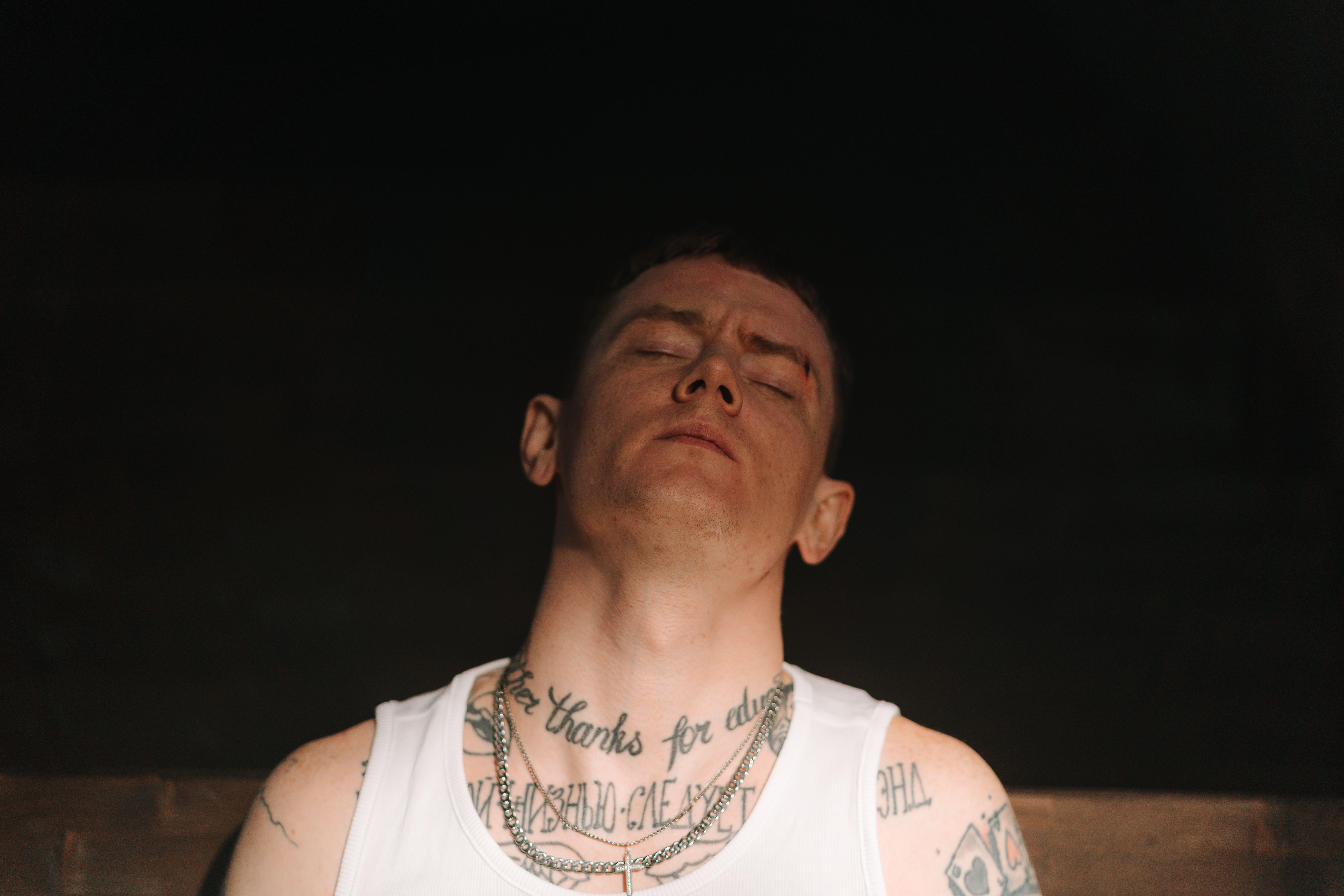 Intimate portrait of a tattooed man with eyes closed, exuding a punk vibe in dim lighting.