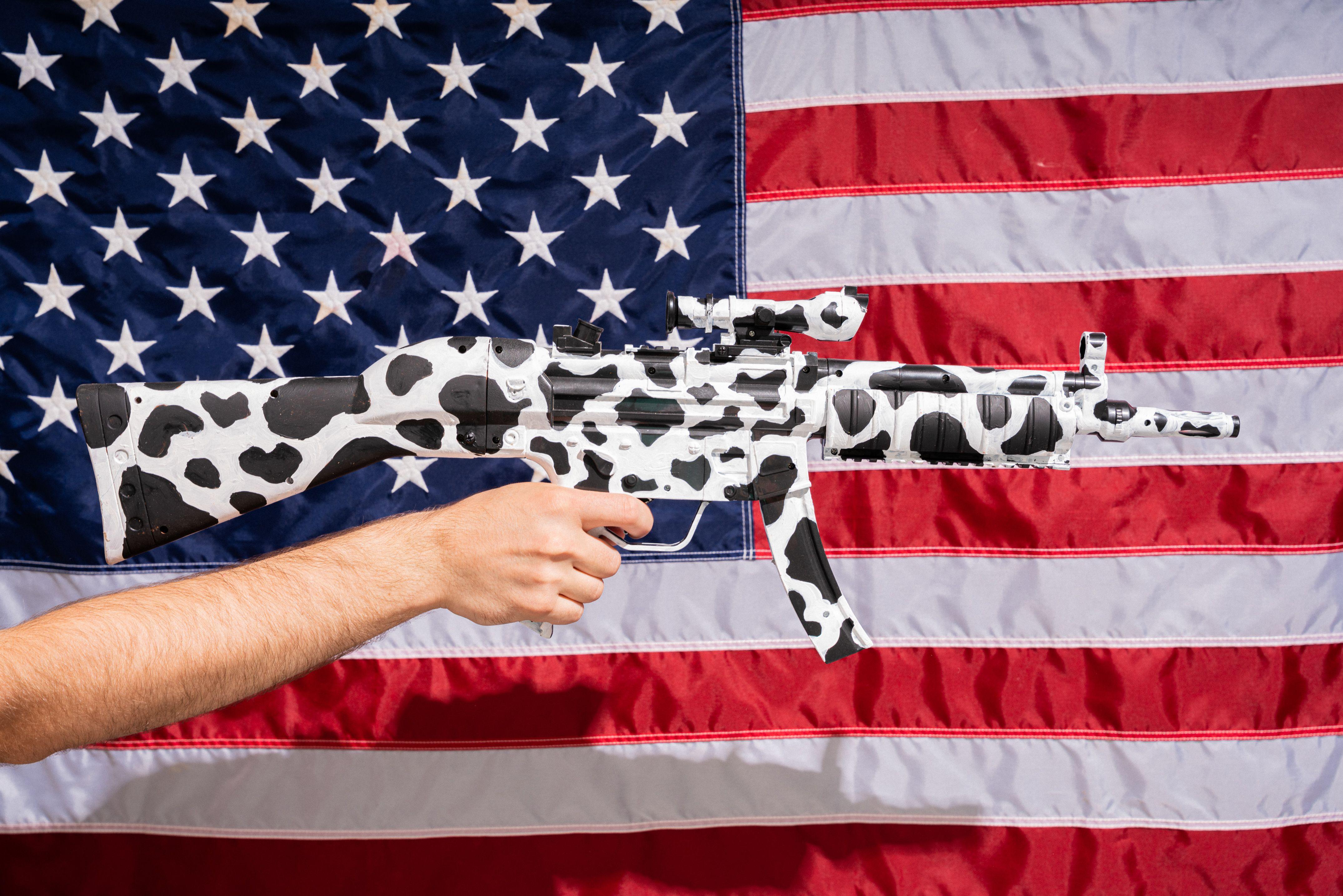 Cow-patterned rifle held in front of American flag, symbolizing culture clash.