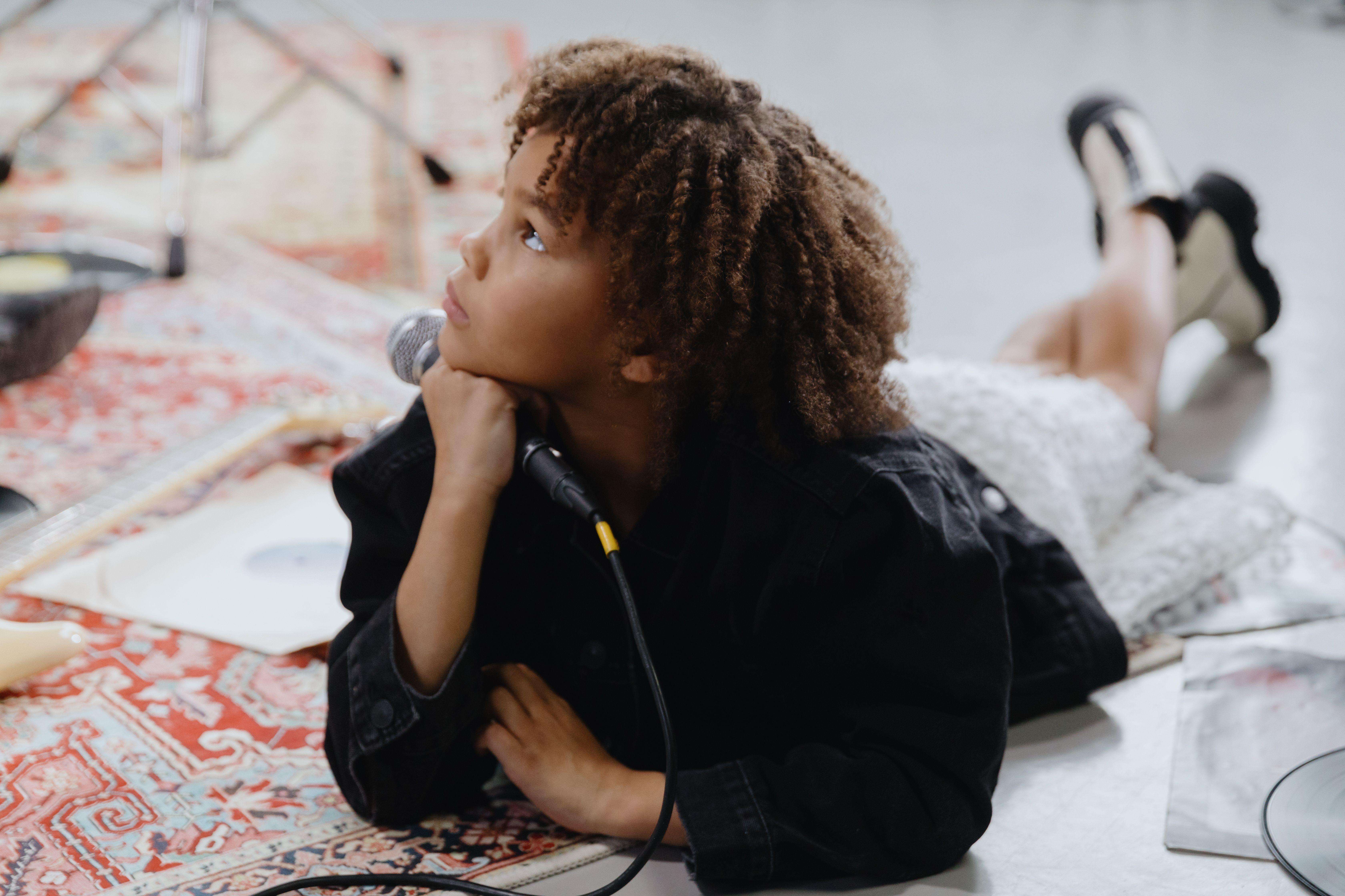 Curly-haired child lies indoors with a microphone, looking thoughtfully upwards.