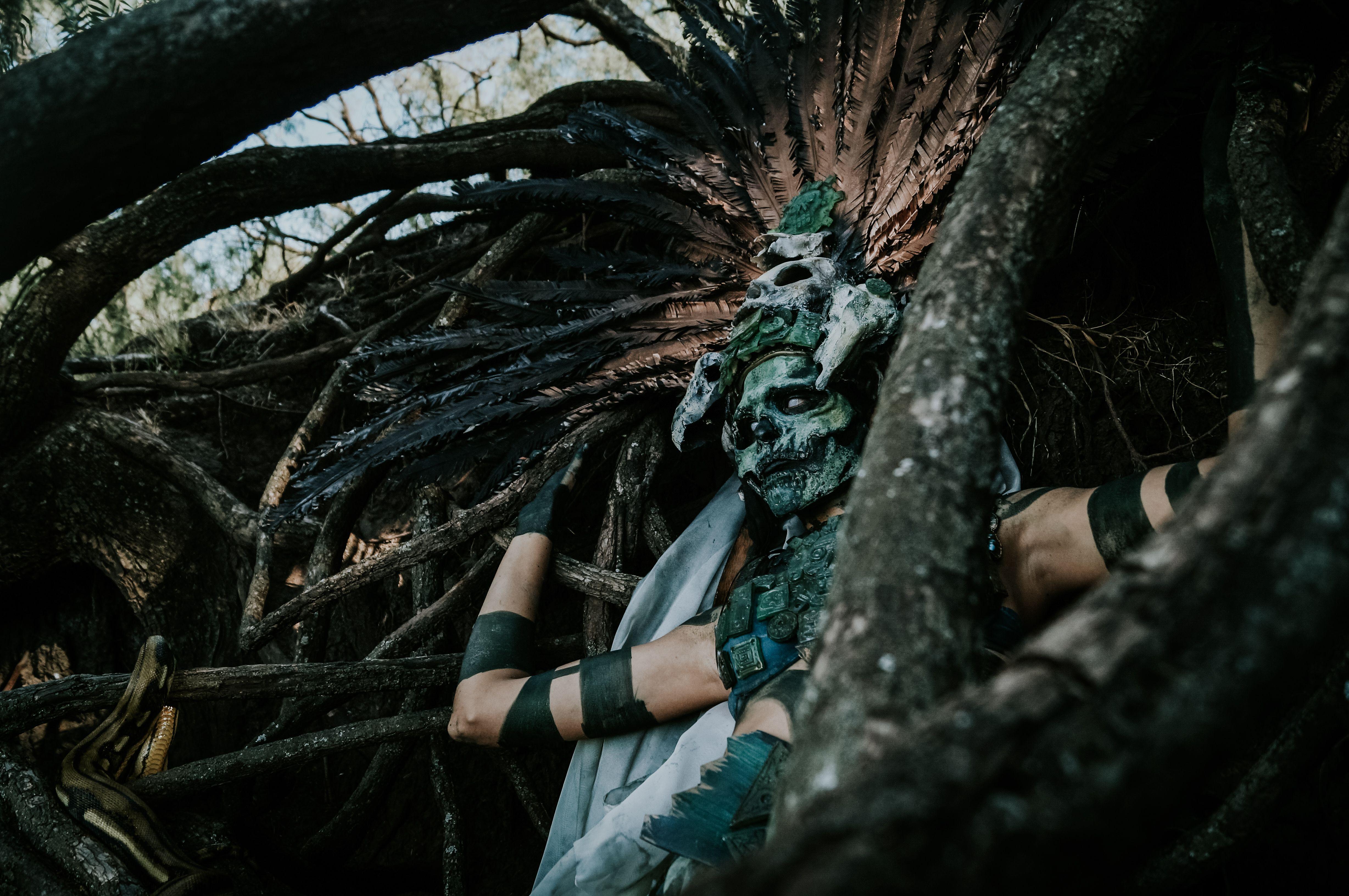 A person in an elaborate headdress and mask blends with twisted tree branches, evoking mythological themes.