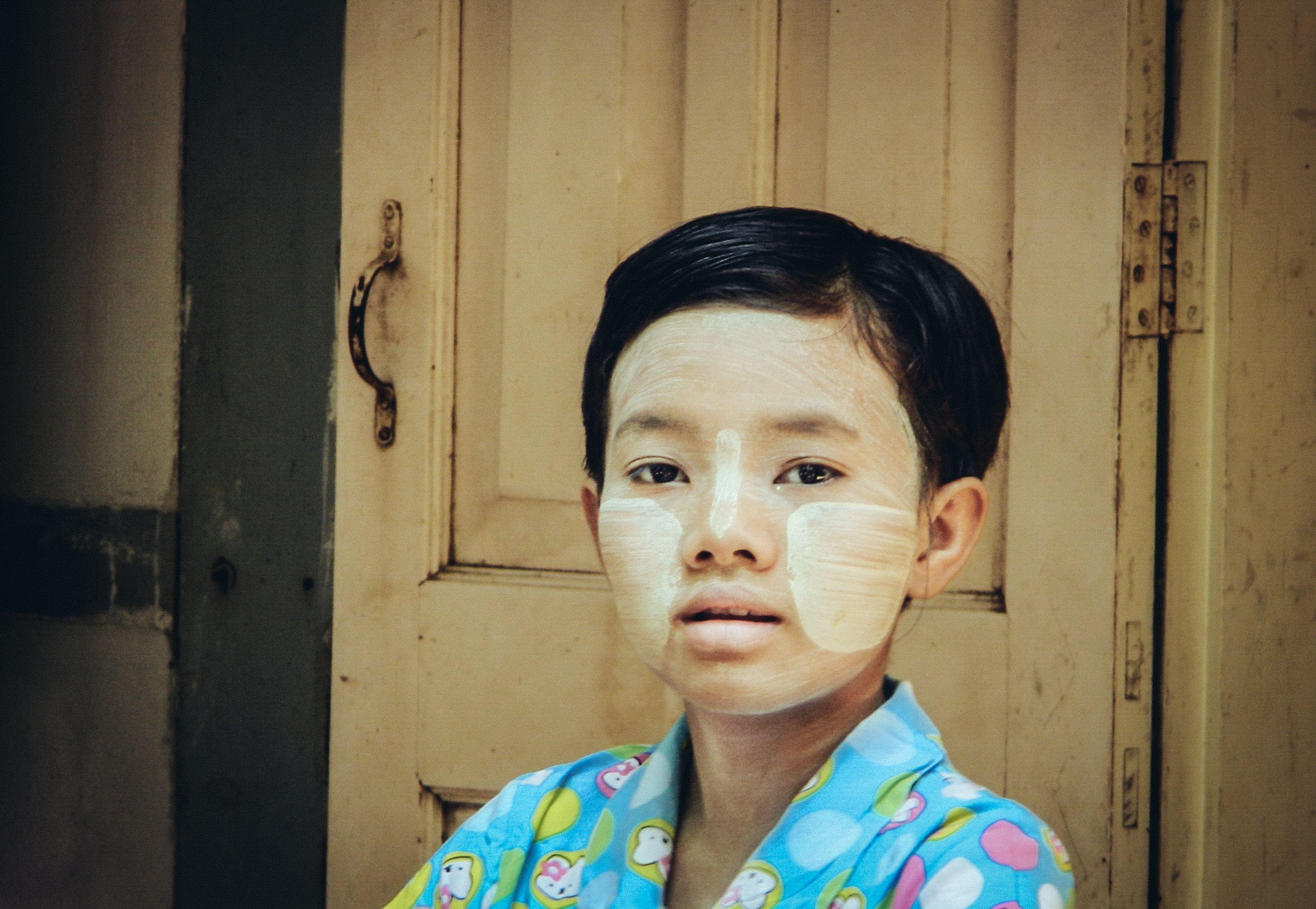 A young child with thanaka on their face, standing in front of an old door in Myanmar.