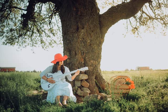 A woman in a red hat plays an acoustic guitar seated under a large tree outdoors.