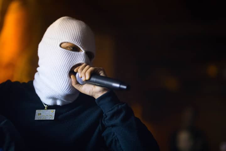 Masked artist wearing a white balaclava performing live on stage with a microphone.