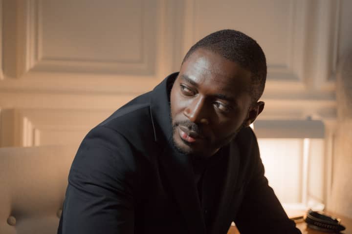 Thoughtful man in a black suit seated in a warmly lit indoor setting, reflecting an elegant mood.