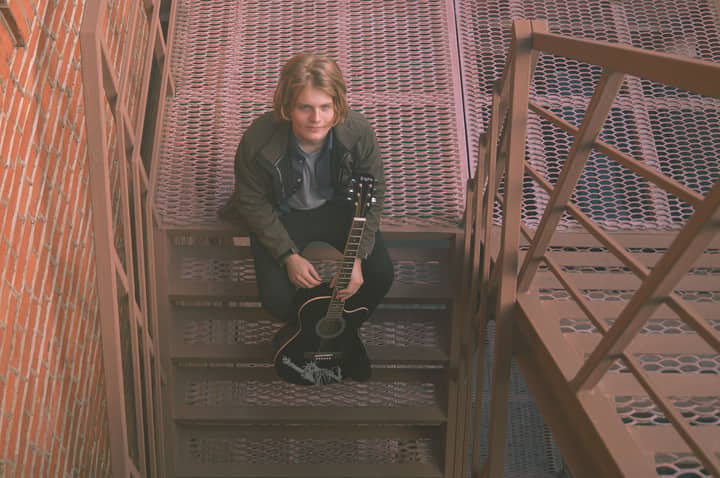 A young man with a guitar sitting on a metal staircase, looking up.