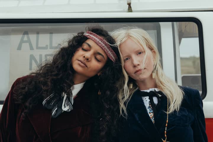 Two stylish women in hippie attire lean against a vintage van, exuding a 60s vibe.