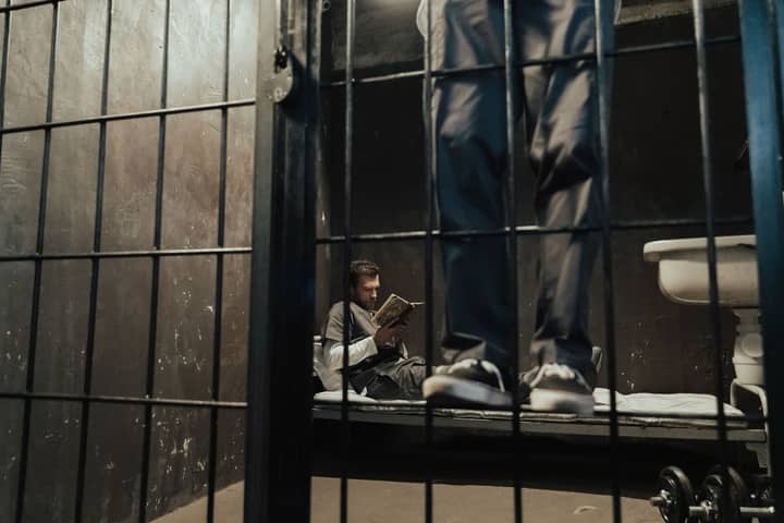 Inmate reading a book inside a prison cell while another stands nearby.
