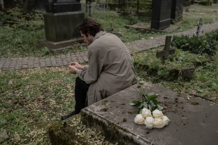 A man in a trench coat sits grieving at a cemetery, white roses on a tombstone.