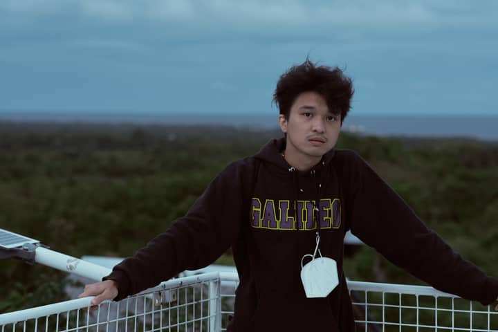 A young man wearing a black hoodie standing outdoors with a scenic background and a face mask hanging around his neck.