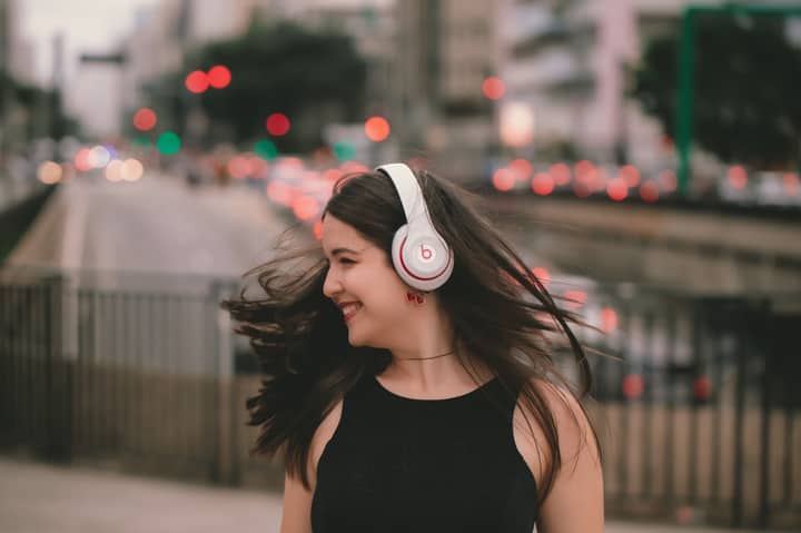 Young woman wearing wireless headphones, smiling joyfully in a city street background, enjoying music.