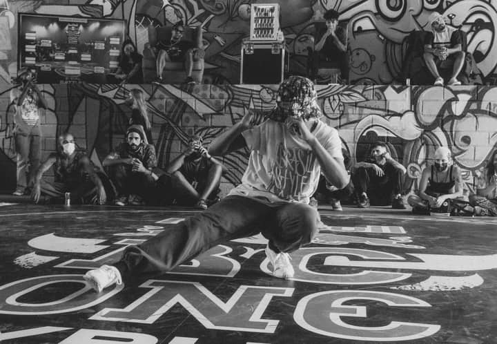 A breakdancer performs energetically in front of a graffiti wall, surrounded by an audience.