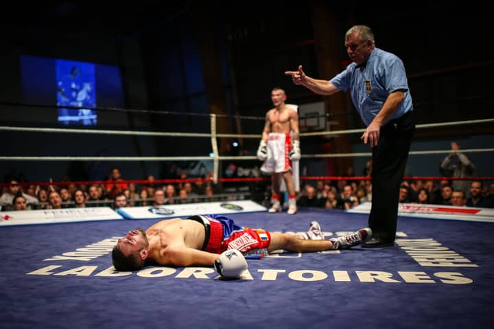 A boxer lies knocked out in the ring as the referee counts, with a crowd watching intently.