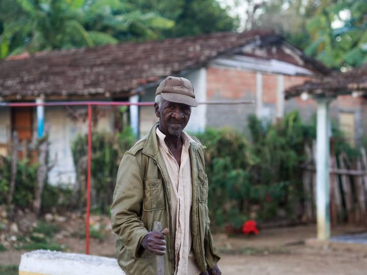 Senior man standing in a rural outdoor scene with rustic buildings and vegetation.