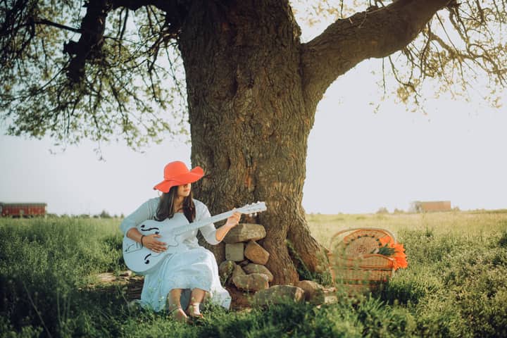 A woman in a red hat plays an acoustic guitar seated under a large tree outdoors.