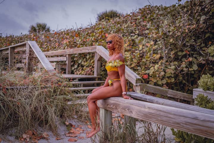 African American woman in yellow bikini sitting on wooden bench outdoors.