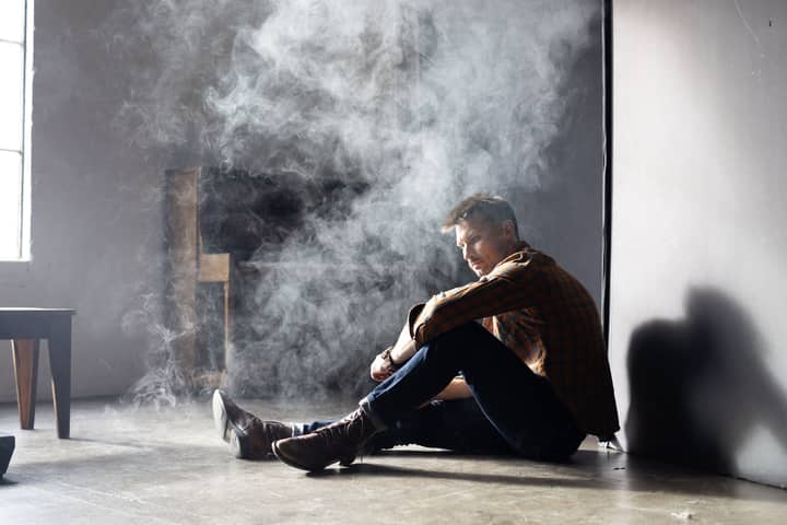 A man in boots and denim sits pensively in a smoke-filled room with soft natural light.