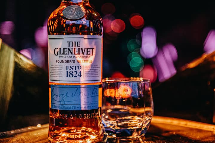 A Glenlivet bottle and glass on a tray with colorful bokeh lights in the background.