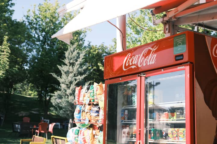 Outdoor Coca-Cola refrigerator beside snacks and trees on a sunny day.