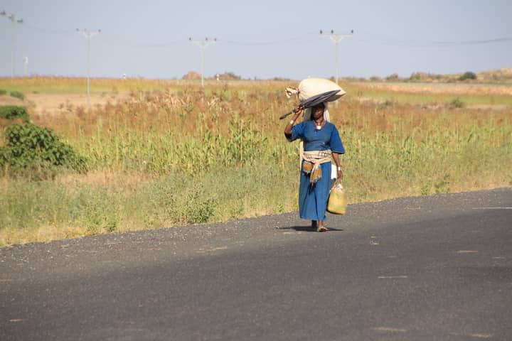 A barefooted Ethiopian woman walks along a rural road carrying goods on her head.