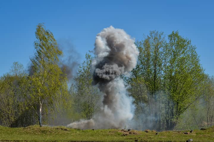 A powerful explosion creates a huge cloud of smoke against a backdrop of green trees, illustrating nature vs. destruction.