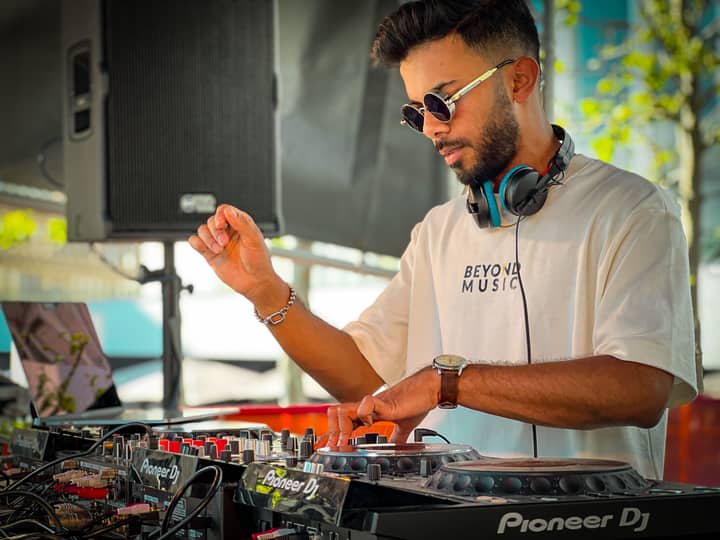 A stylish DJ wearing sunglasses and headphones mixes tracks outdoors using a turntable.
