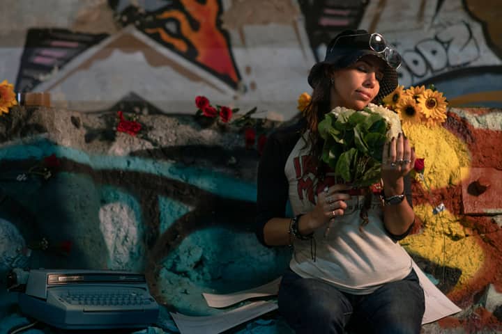 A woman in a bucket hat holds a bouquet, blending nature with urban graffiti art in Mexico City.