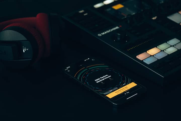 Dark-themed music setup featuring a DJ controller, smartphone, and headphones on a sleek surface.