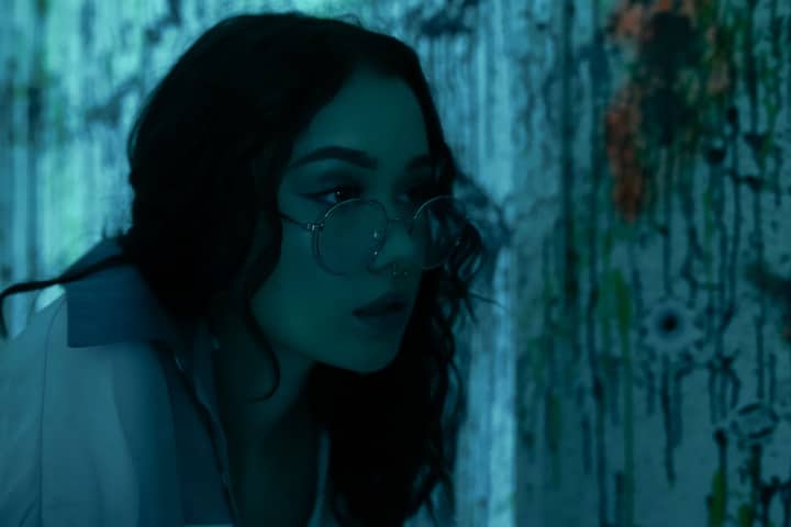 Close-up artistic portrait of a woman with glasses and nose ring in a modern paint-splattered room.