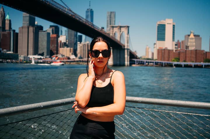Woman in black tank top and sunglasses posing with the Brooklyn Bridge in the background.