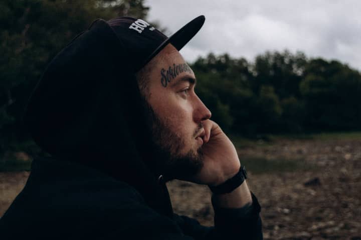 A thoughtful bearded man with a cap and visible tattoo in a natural outdoor setting.