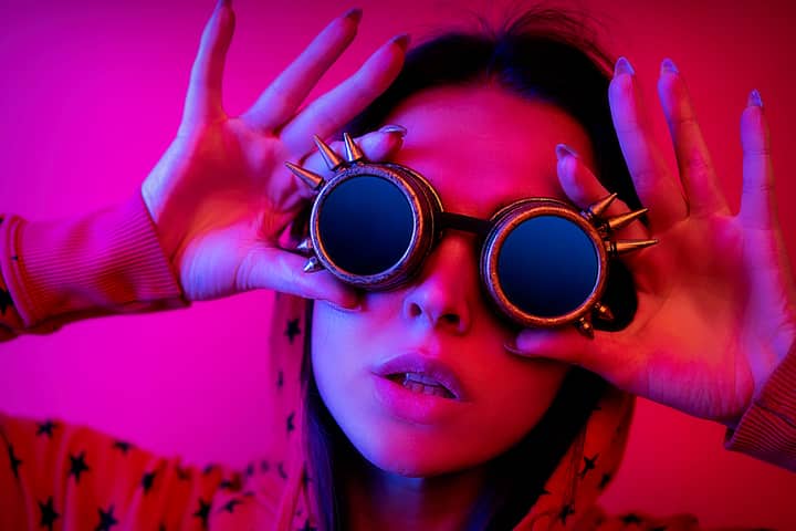 Vibrant and edgy portrait of a woman wearing spiked sunglasses and a pink hoodie against a monochrome backdrop.