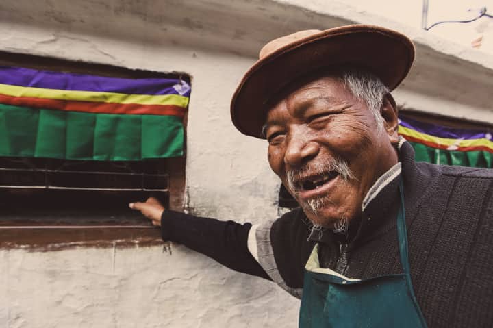 A happy elderly man with a hat and mustache smiling by a colorful window.