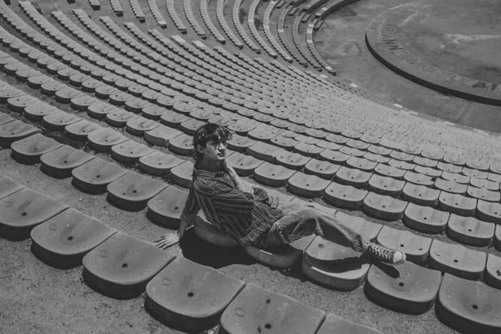 A man sits alone in empty stadium seating, captured in a monochrome shot.